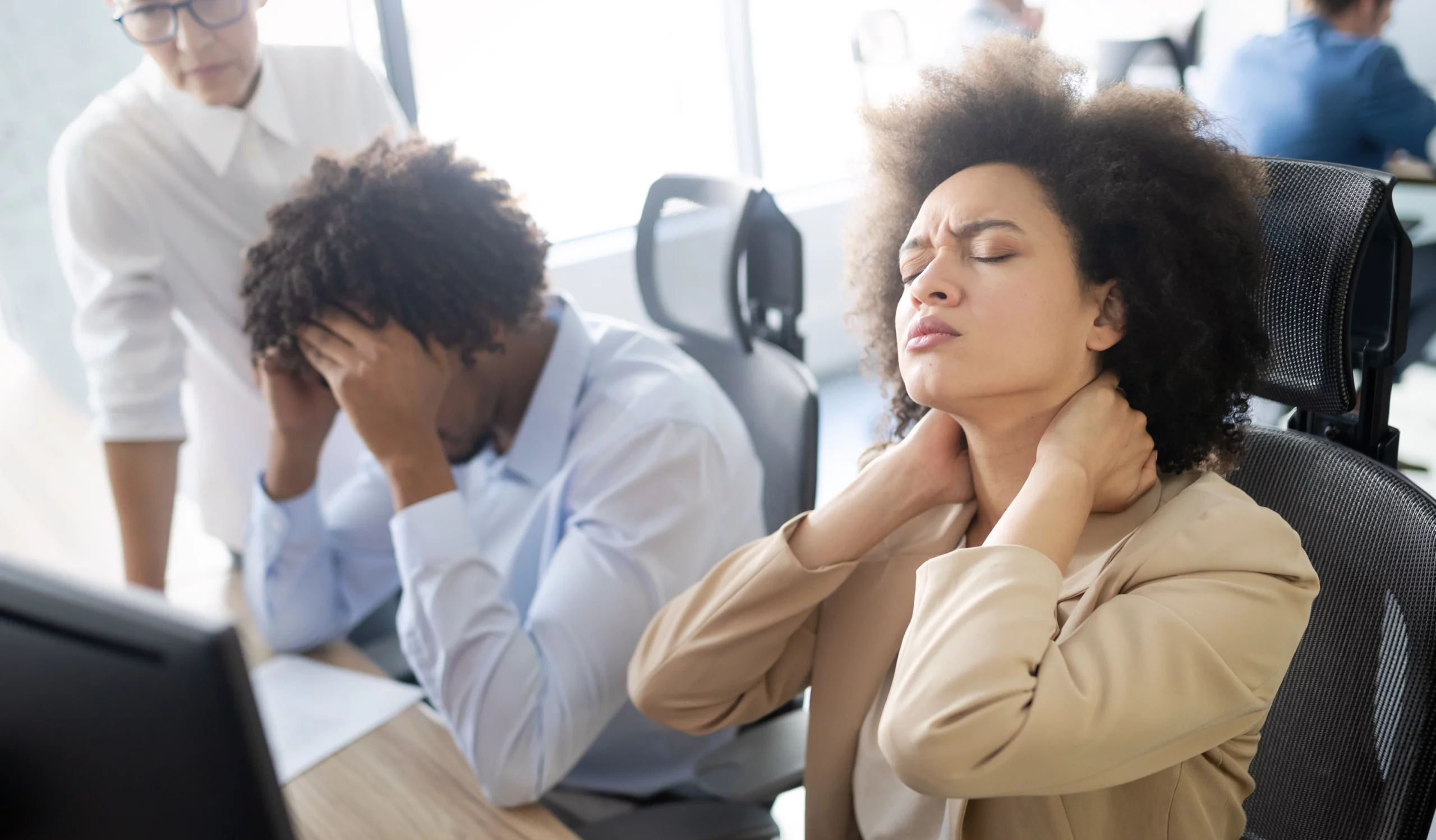 Three office workers in a bright room, two seated, showing stress. A woman massages her neck, while a man covers his face. Another stands, observing.